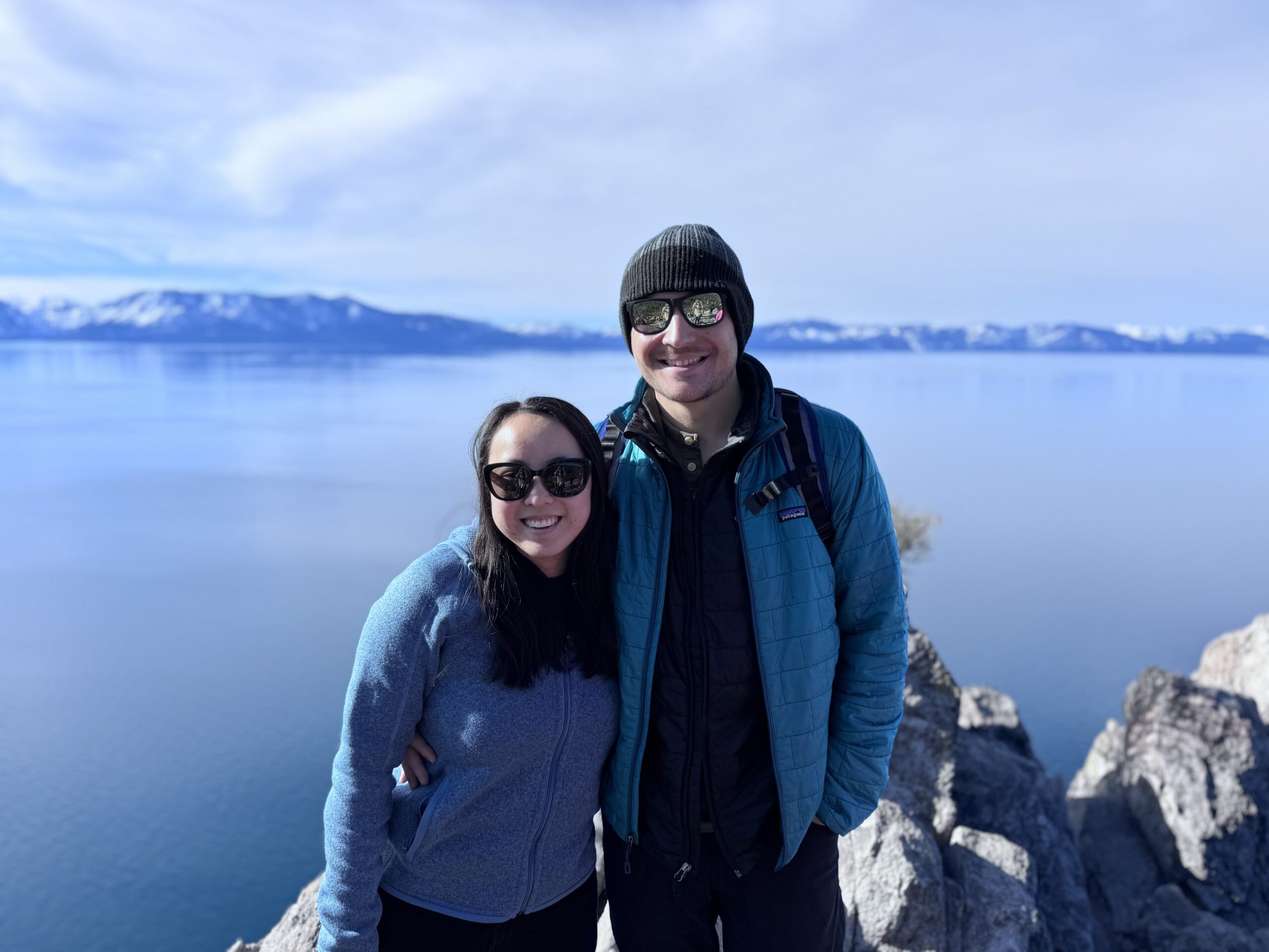 Mary and James standing on a rock overlooking Lake Tahoe and snow capped mountains.